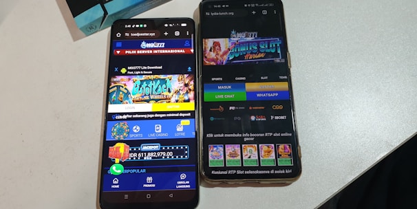 Two smartphones are displayed on a white surface, both showing gambling websites with colorful graphics, text, and various options for logging in and accessing games. The screens are bright and filled with promotional content related to casino and slot games.